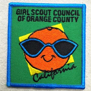 May include: A square embroidered patch with a green background and blue border. It features an orange circle wearing blue-rimmed black sunglasses, a yellow square behind it, and the text Girl Scout Council of Orange County and California.