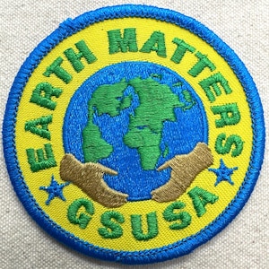 May include: Embroidered patch with the words "EARTH MATTERS" and "GSUSA" in green and yellow. The patch features a blue globe with green continents, brown hands, and blue stars. The patch is round with a blue border.