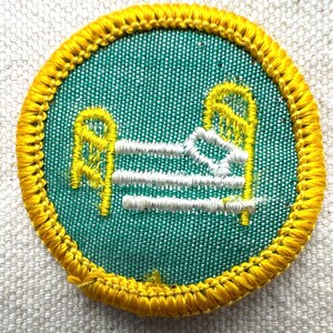 May include: A round patch with a green background and a yellow border. The patch features a bed design in yellow and white thread. The bed has a headboard, footboard, and three horizontal lines representing the mattress.
