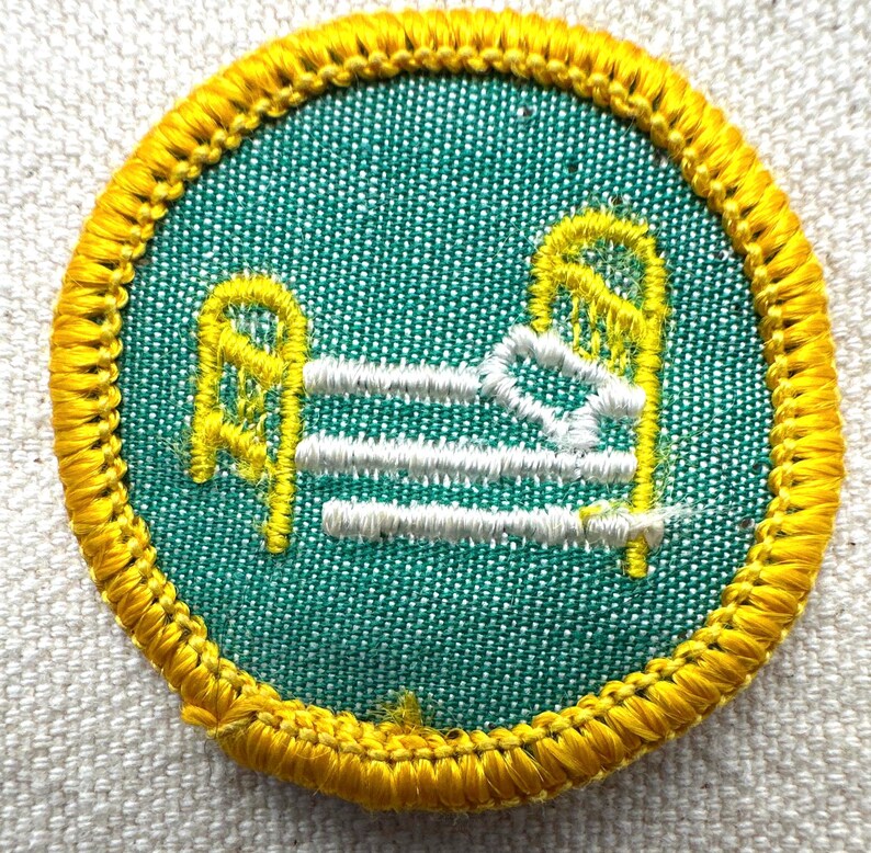 May include: A round patch with a green background and a yellow border. The patch features a bed design in yellow and white thread. The bed has a headboard, footboard, and three horizontal lines representing the mattress.