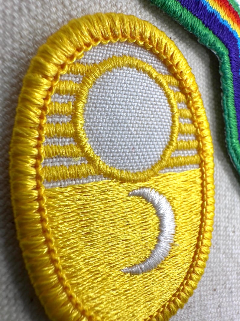 Puede incluir: Parche amarillo bordado con un dise&ntilde;o de luna creciente blanca y sol. El parche tiene un borde amarillo y est&aacute; sobre una tela gris claro. El dise&ntilde;o es un tema celestial estilizado.