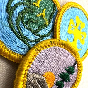 May include: Close-up of three embroidered patches with yellow borders. One patch features blue waves and green details with yellow birds. Another has a yellow tree on a blue background. The third patch shows a mountain, sun, and trees.
