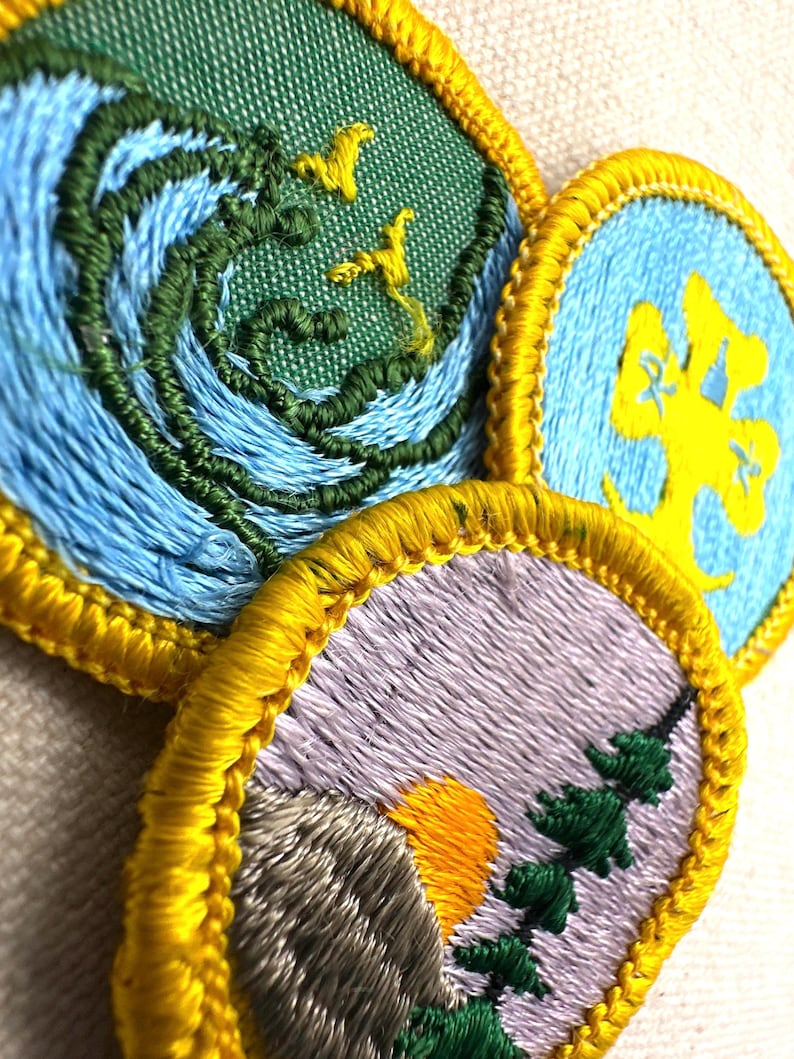 May include: Close-up of three embroidered patches with yellow borders. One patch features blue waves and green details with yellow birds. Another has a yellow tree on a blue background. The third patch shows a mountain, sun, and trees.