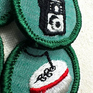 May include: Close-up of embroidered patches with green borders. One patch features a black and white walkie-talkie design. Another patch shows a white shoe with a red sole and black detailing. The patches are on a textured, light-colored background.