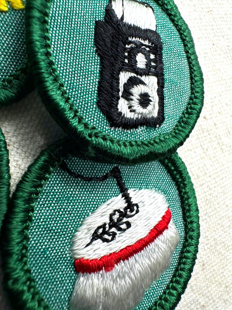 May include: Close-up of embroidered patches with green borders. One patch features a black and white walkie-talkie design. Another patch shows a white shoe with a red sole and black detailing. The patches are on a textured, light-colored background.