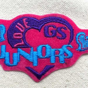 Puede incluir: Un parche de fieltro rosa con texto bordado y dise&ntilde;os de coraz&oacute;n en azul y morado. El parche dice "LOVE GS JUNIORS" con una forma de coraz&oacute;n en el centro. Dos corazones azules est&aacute;n a cada lado del parche.