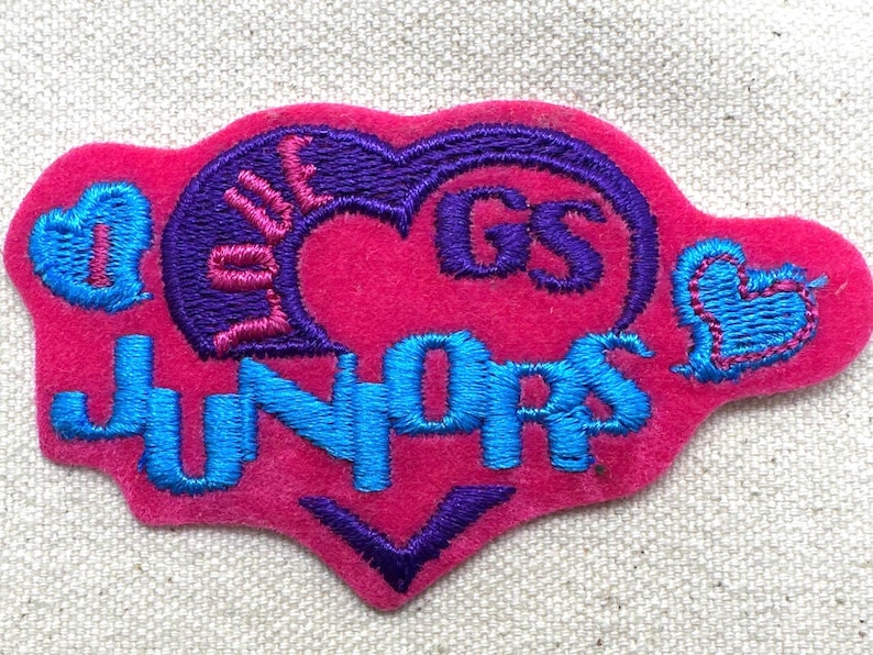 Puede incluir: Un parche de fieltro rosa con texto bordado y dise&ntilde;os de coraz&oacute;n en azul y morado. El parche dice "LOVE GS JUNIORS" con una forma de coraz&oacute;n en el centro. Dos corazones azules est&aacute;n a cada lado del parche.