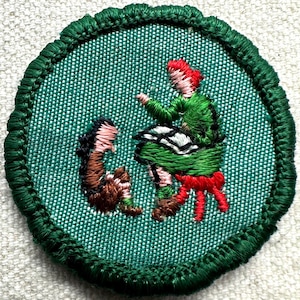 May include: Embroidered patch with a green background and a green border. The patch features a figure in a green outfit with red hair and a red sash, and a brown figure. The figures appear to be dancing.