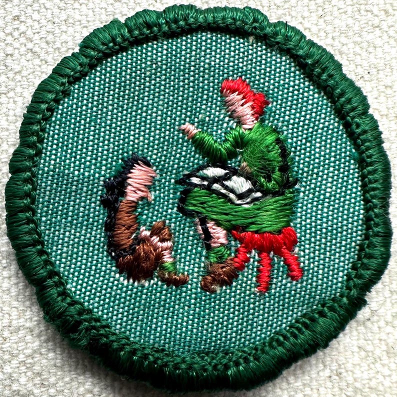 May include: Embroidered patch with a green background and a green border. The patch features a figure in a green outfit with red hair and a red sash, and a brown figure. The figures appear to be dancing.