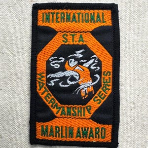 May include: Rectangular patch with black border, orange and green text, and a central emblem. The text reads "INTERNATIONAL S.T.A. WATERMANSHIP SERIES MARLIN AWARD." The emblem features a marlin in orange and white.