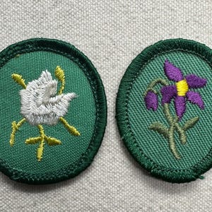 May include: Two embroidered patches with green borders. One features a white dove with yellow accents, the other a purple flower with a yellow center. Both are on a green background.