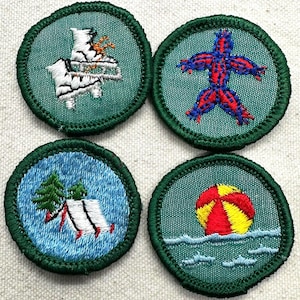 May include: Four embroidered patches with green borders. The patches feature various designs: a polar bear on a sled, a stick figure, a tent with trees, and a beach ball in the water. Each patch is round and has a textured background.