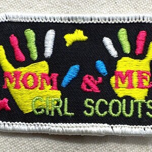 May include: Embroidered patch with a black background and white border. The patch features two yellow handprints with the words "MOM & ME" in pink and yellow. The words "GIRL SCOUTS" are in green.