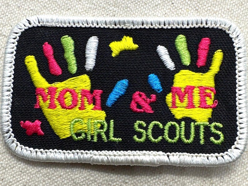 May include: Embroidered patch with a black background and white border. The patch features two yellow handprints with the words "MOM & ME" in pink and yellow. The words "GIRL SCOUTS" are in green.