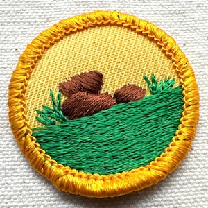 May include: Embroidered patch with a yellow background and a detailed design. The patch features brown rocks, green grass, and a yellow border. The design is stitched with various colors of thread, creating a textured appearance.