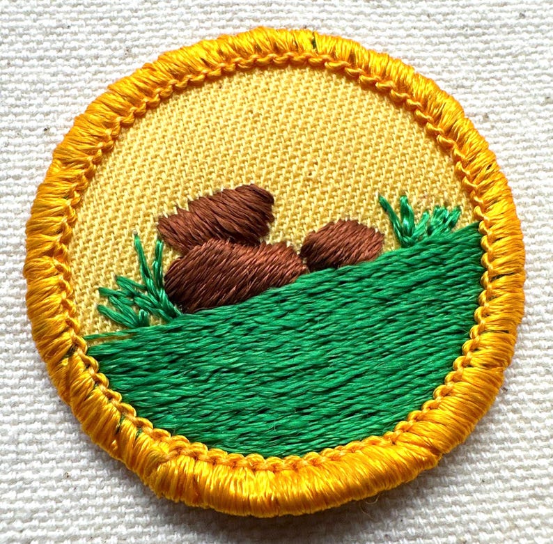 May include: Embroidered patch with a yellow background and a detailed design. The patch features brown rocks, green grass, and a yellow border. The design is stitched with various colors of thread, creating a textured appearance.