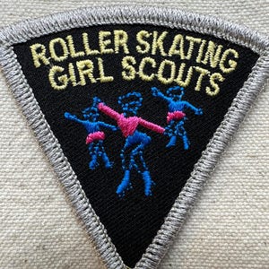 Puede incluir: Un parche triangular negro con las palabras "ROLLER SKATING GIRL SCOUTS" bordadas en oro. El parche presenta tres figuras en azul y rosa, con un borde plateado.