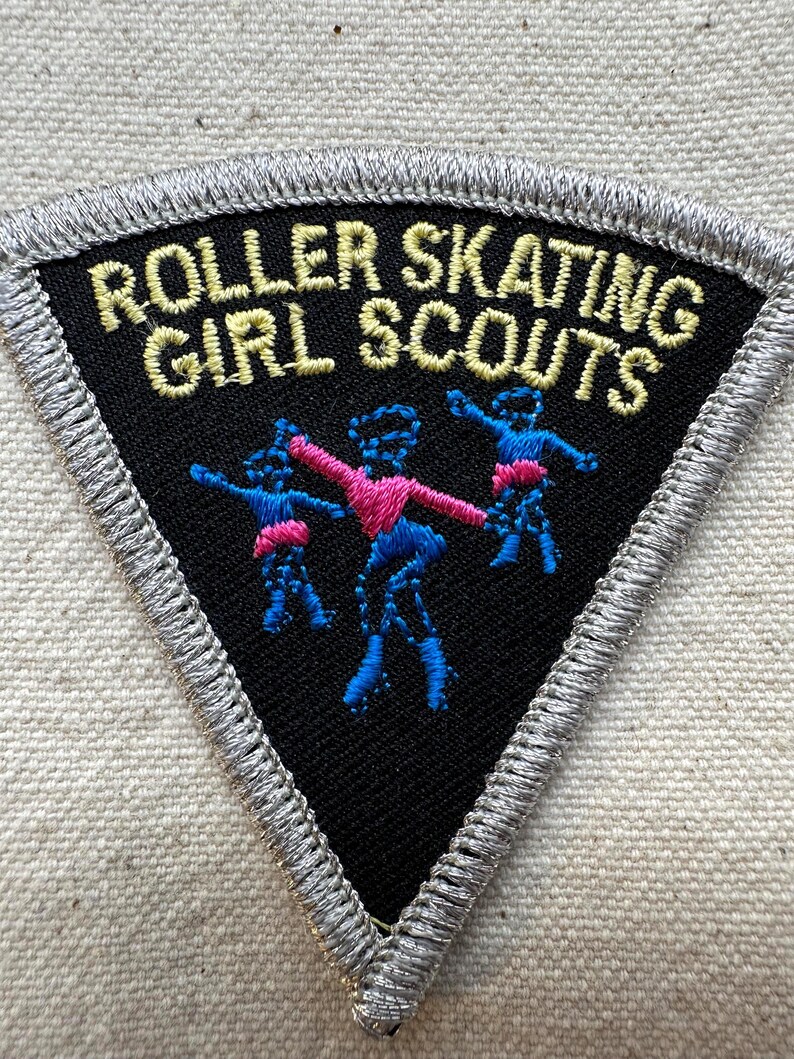 Puede incluir: Un parche triangular negro con las palabras "ROLLER SKATING GIRL SCOUTS" bordadas en oro. El parche presenta tres figuras en azul y rosa, con un borde plateado.