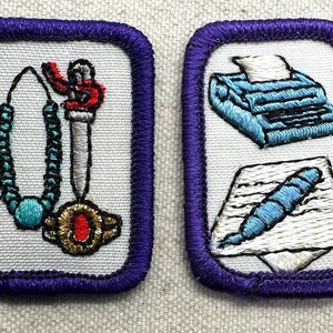 May include: Two embroidered patches with purple borders. One features a turquoise beaded necklace and a pendant with a red accent. The other shows a blue typewriter and a pen on paper. Both are on a white background.