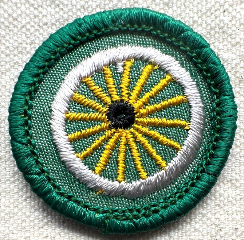 May include: Embroidered patch with a circular design. The patch features a green outer border, a white inner ring, and a central design of yellow spokes radiating from a black center. The background is a light green fabric.