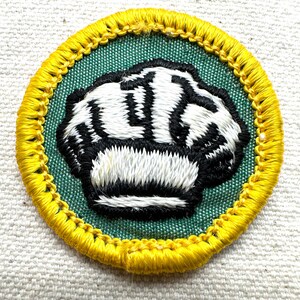 May include: Embroidered patch featuring a clenched fist design. The patch has a green center with a black and white fist, surrounded by a yellow border. The patch is circular and appears to be made of fabric.