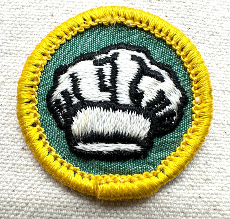 May include: Embroidered patch featuring a clenched fist design. The patch has a green center with a black and white fist, surrounded by a yellow border. The patch is circular and appears to be made of fabric.