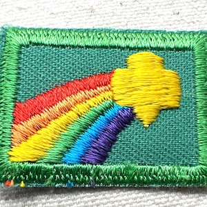 May include: Embroidered patch featuring a rainbow and a yellow emblem on a green background. The patch has a green border and is rectangular in shape. The rainbow arcs from red to purple.
