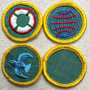 May include: Four embroidered patches with a green background and yellow border. Designs include a life preserver, a globe, a blue bird, and a blank patch. The patches are circular and likely for a uniform or craft project.