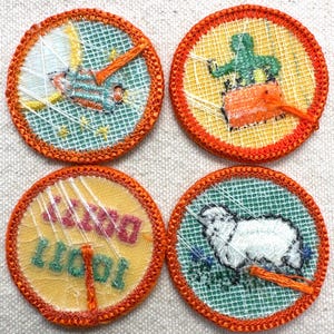 May include: Four round embroidered patches with orange borders. Each patch features a different design: a bird, a figure with a long nose, text that reads "Doing Better", and a sheep. The backgrounds are yellow, teal, and white.