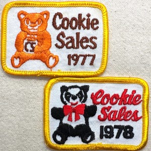 May include: Two embroidered patches with a white background and gold trim. One features an orange bear and the text "Cookie Sales 1977." The other shows a panda bear and the text "Cookie Sales 1978."