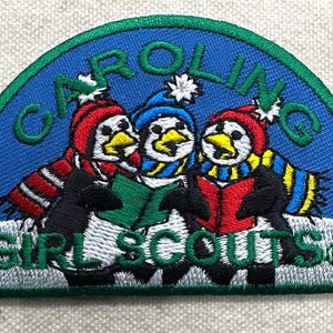 May include: Embroidered patch featuring three penguins wearing winter hats and scarves, holding a book. The patch has a blue background with the words "CAROLING" and "GIRL SCOUTS" in green.