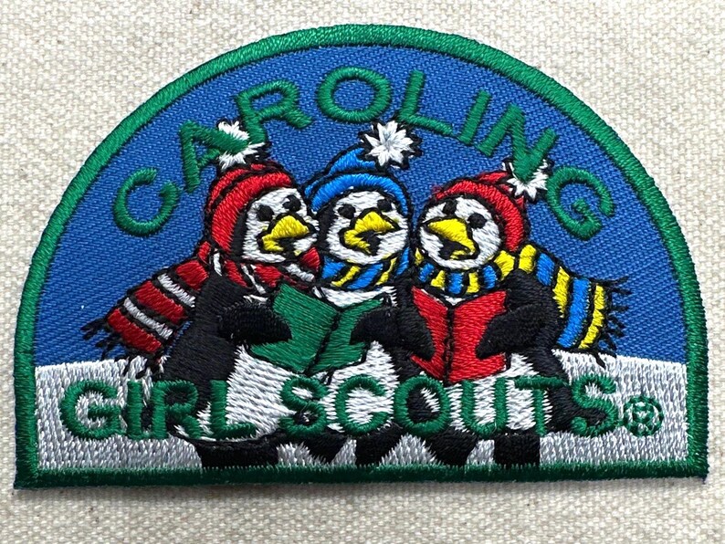 May include: Embroidered patch featuring three penguins wearing winter hats and scarves, holding a book. The patch has a blue background with the words "CAROLING" and "GIRL SCOUTS" in green.