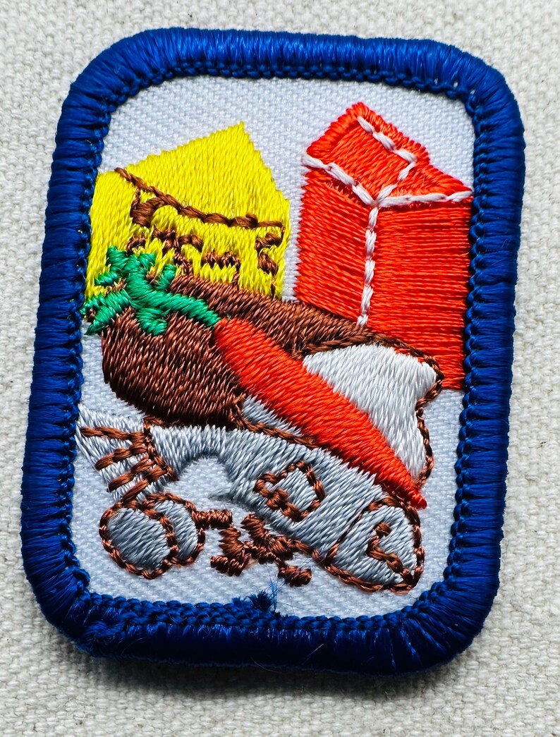 May include: Embroidered patch with a blue border. The patch features a brown bird with an orange beak, a yellow house, and a red gift box. The bird is perched on a gray object with the word "One" embroidered on it.