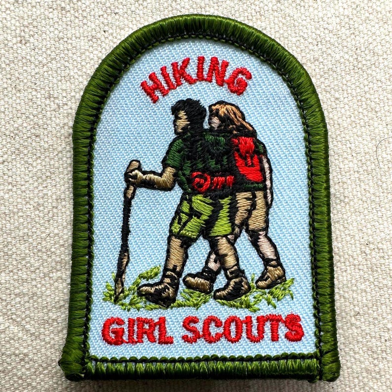 May include: A light blue patch with the words "HIKING" and "GIRL SCOUTS" in red. The patch features two figures hiking with a walking stick and a red backpack, outlined in green.