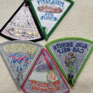 Puede incluir: Una colecci&oacute;n de cinco parches triangulares bordados con varios dise&ntilde;os y texto. Los parches presentan diferentes colores, incluyendo verde, rosa y azul, con fondos blancos y grises. El texto en los parches est&aacute; en varias fuentes y colores.
