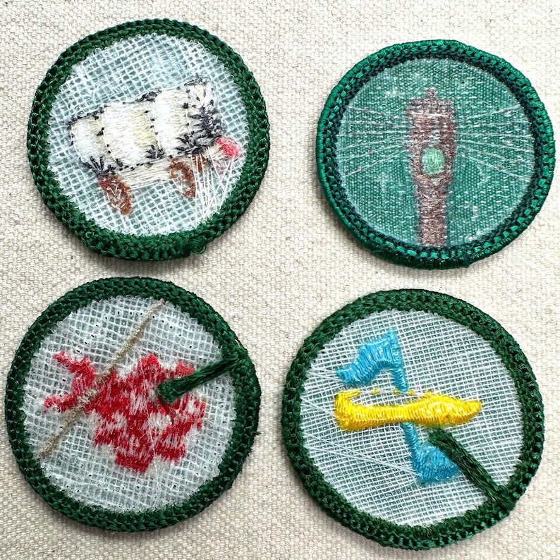 May include: Four round embroidered patches with green borders. The patches feature images of a sheep, a lighthouse, a red flower, and a yellow and blue design. The patches are on a light-colored fabric.