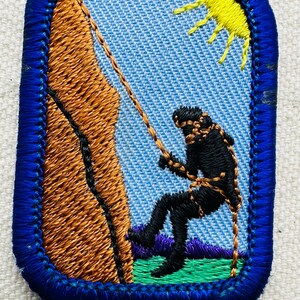 May include: Embroidered patch featuring a rock climber rappelling down a brown rock face. The climber is black, with a rope and harness. A yellow sun is in the sky. The patch has a blue border and a light blue background.