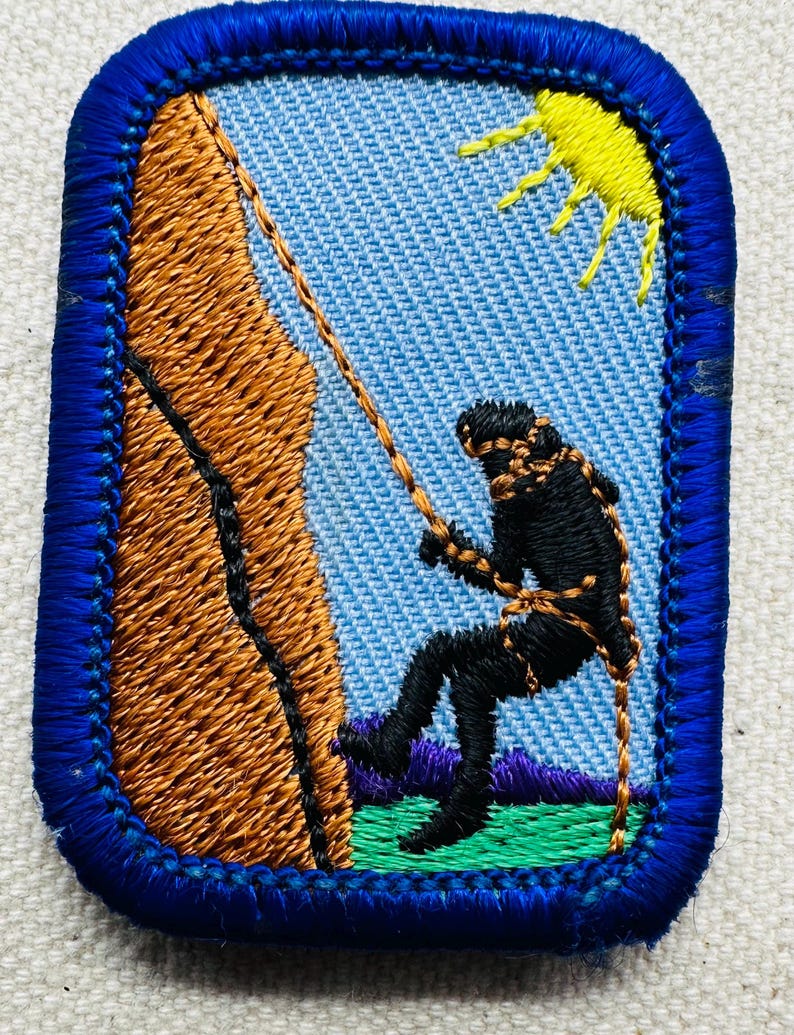 May include: Embroidered patch featuring a rock climber rappelling down a brown rock face. The climber is black, with a rope and harness. A yellow sun is in the sky. The patch has a blue border and a light blue background.