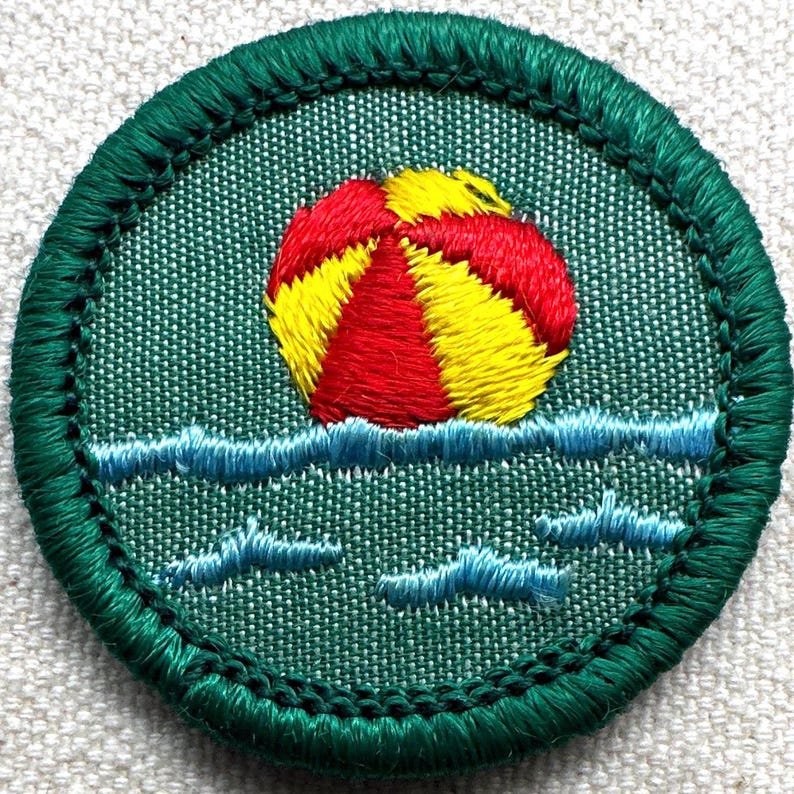 May include: Embroidered patch featuring a red and yellow beach ball floating on blue waves. The patch is round with a green border and a green background. The design evokes a summer theme.