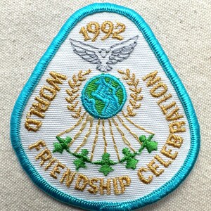 May include: Embroidered patch with a teal border and white background. The patch features the text "WORLD FRIENDSHIP CELEBRATION" and "1992". A globe, dove, and figures holding hands are also embroidered on the patch.