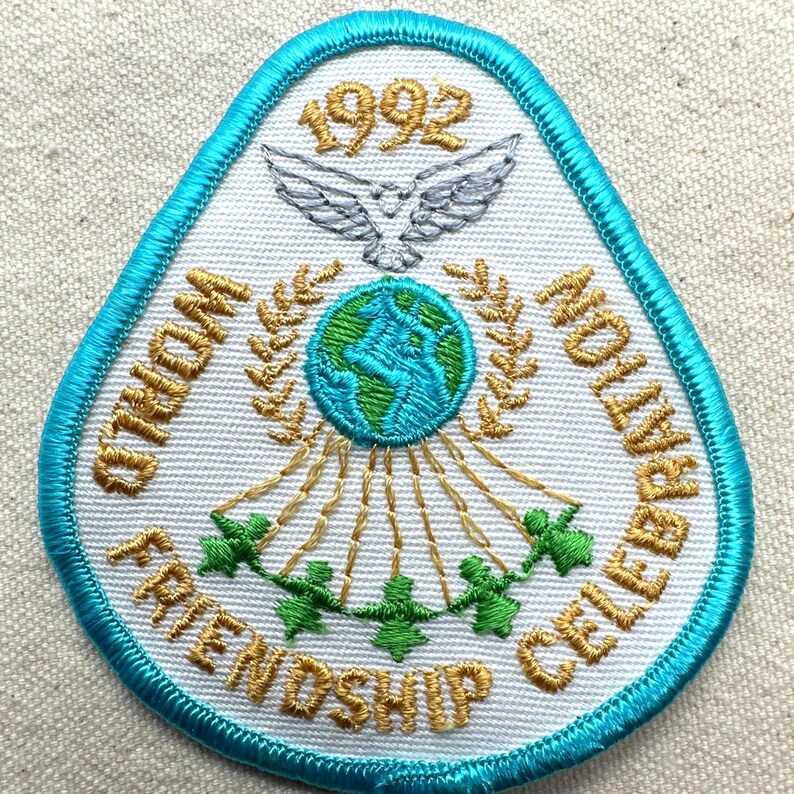 May include: Embroidered patch with a teal border and white background. The patch features the text "WORLD FRIENDSHIP CELEBRATION" and "1992". A globe, dove, and figures holding hands are also embroidered on the patch.