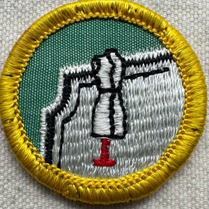 May include: Embroidered patch with a yellow border, green background, and a central design featuring a dressmaker's form. The form is white with black outlines, and the base is red. The patch is circular.