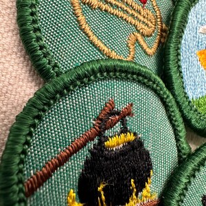 Puede incluir: Primer plano de varios parches bordados con bordes verdes. Un parche presenta una olla sobre un fuego, otro representa un arco y una flecha. Los parches est&aacute;n sobre una tela verde texturizada.