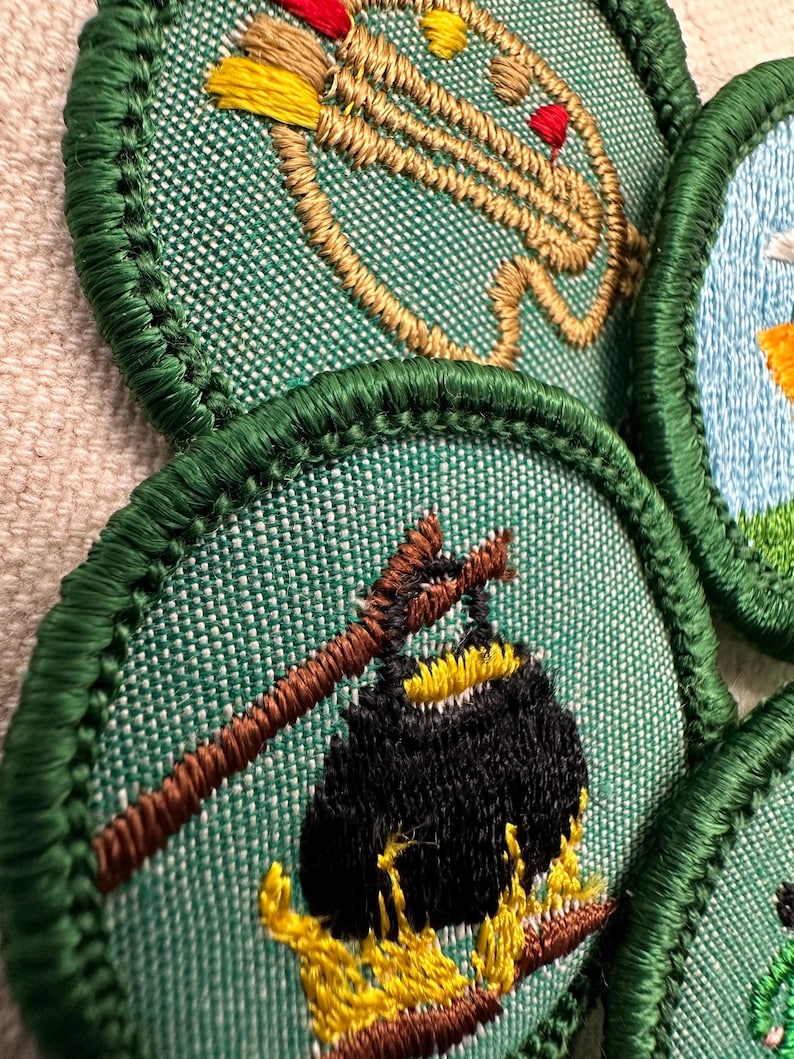 Puede incluir: Primer plano de varios parches bordados con bordes verdes. Un parche presenta una olla sobre un fuego, otro representa un arco y una flecha. Los parches est&aacute;n sobre una tela verde texturizada.
