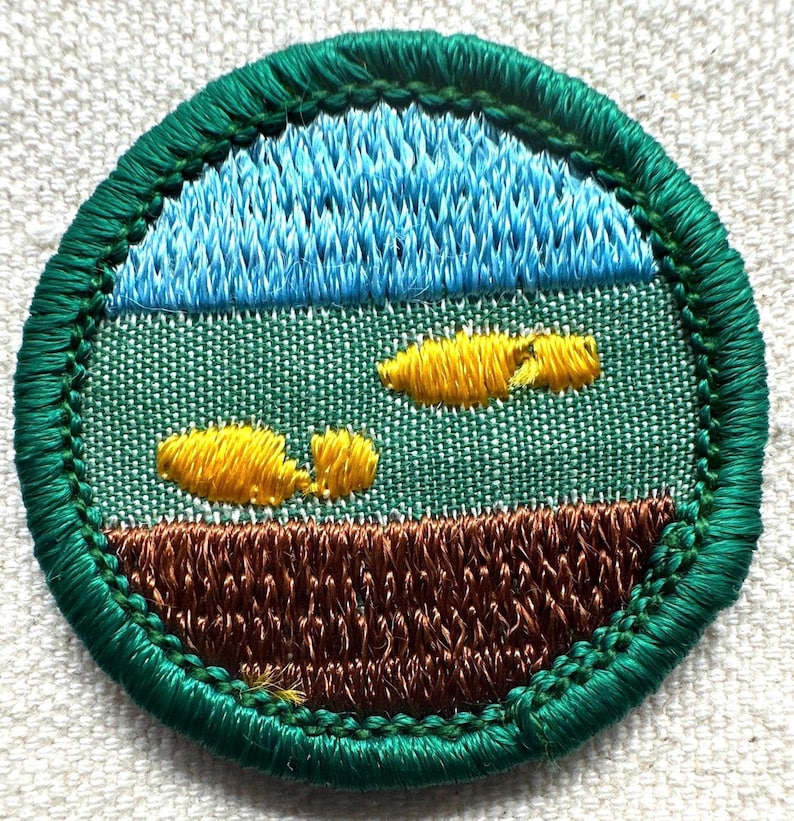 May include: Embroidered patch with a landscape design. The top section features blue stitching representing the sky, followed by a green field with yellow accents. The bottom section is brown, and the patch is bordered with green stitching.