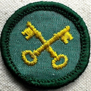 May include: A round, green embroidered patch featuring two crossed yellow keys. The keys are detailed with intricate stitching, set against a textured green background. The patch is bordered with a dark green, stitched edge.