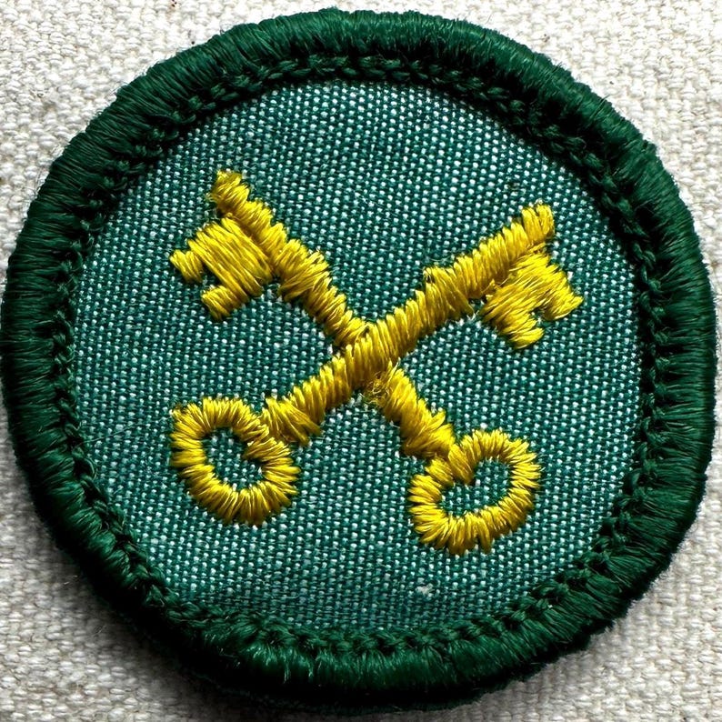May include: A round, green embroidered patch featuring two crossed yellow keys. The keys are detailed with intricate stitching, set against a textured green background. The patch is bordered with a dark green, stitched edge.