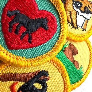May include: Close-up of several embroidered patches with yellow borders. One patch features a black horse inside a red heart on a green background. Other patches depict a raccoon, a campfire, and a brown log.
