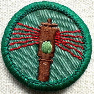 May include: Embroidered patch featuring a brown lighthouse with red light beams radiating outward. The lighthouse has a green accent and is set against a green background. The patch is circular with a green border.