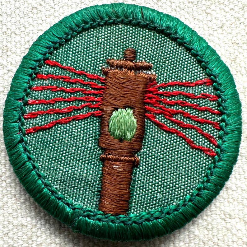 May include: Embroidered patch featuring a brown lighthouse with red light beams radiating outward. The lighthouse has a green accent and is set against a green background. The patch is circular with a green border.